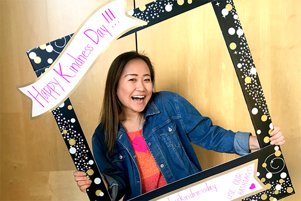 A smiling Cho holds and peeks through a decorative black, gold and pink cardboard frame that reads "Happy Kindness Day" and "Use our Hashtags"